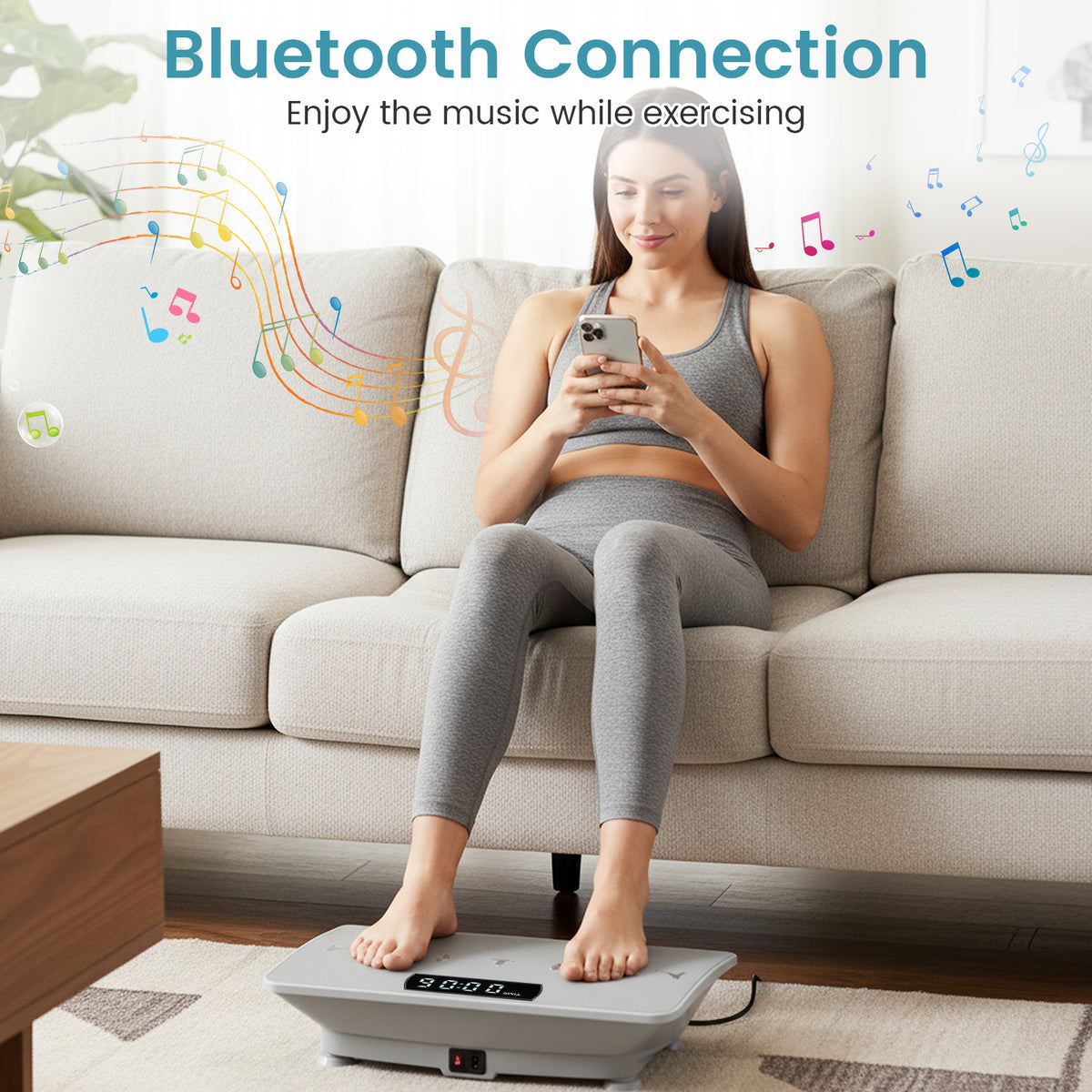 A woman sitting on a sofa using the grey vibration plate machine, connected via Bluetooth