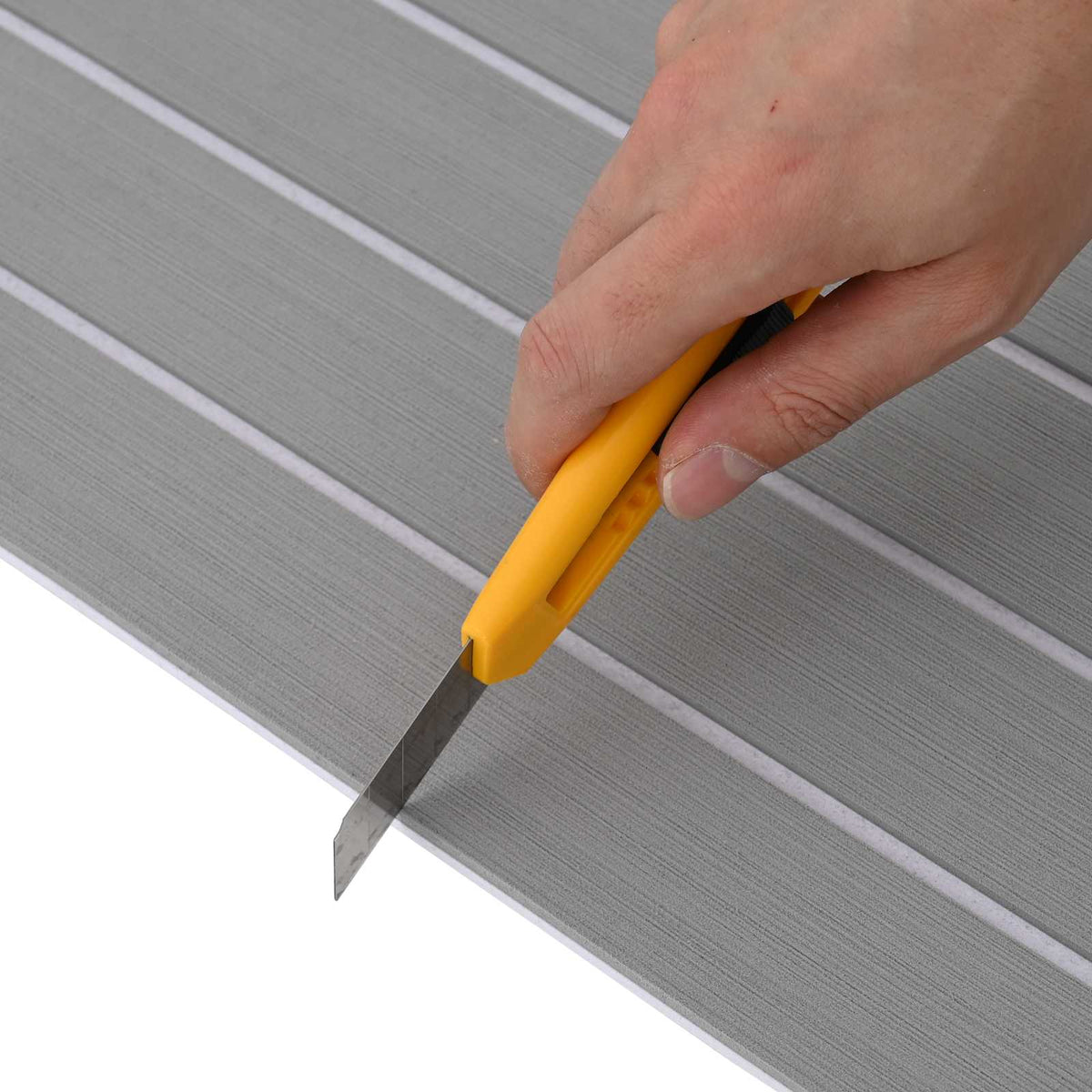 A hand using a yellow utility knife to cut the light grey and white striped boat mat