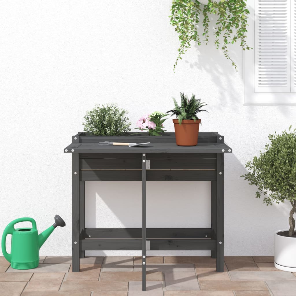 A grey wooden garden planter with a fold-out table set up on a patio with plants and a watering can