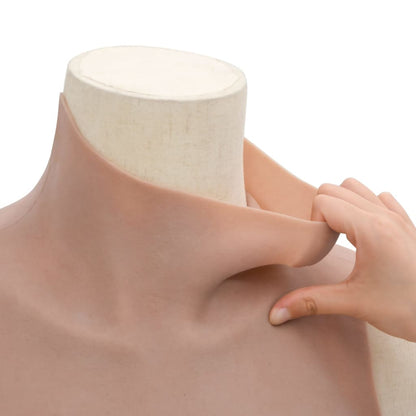 A hand pulls at the neckline of the tan silicone chest piece with D cup breasts, showing flexibility