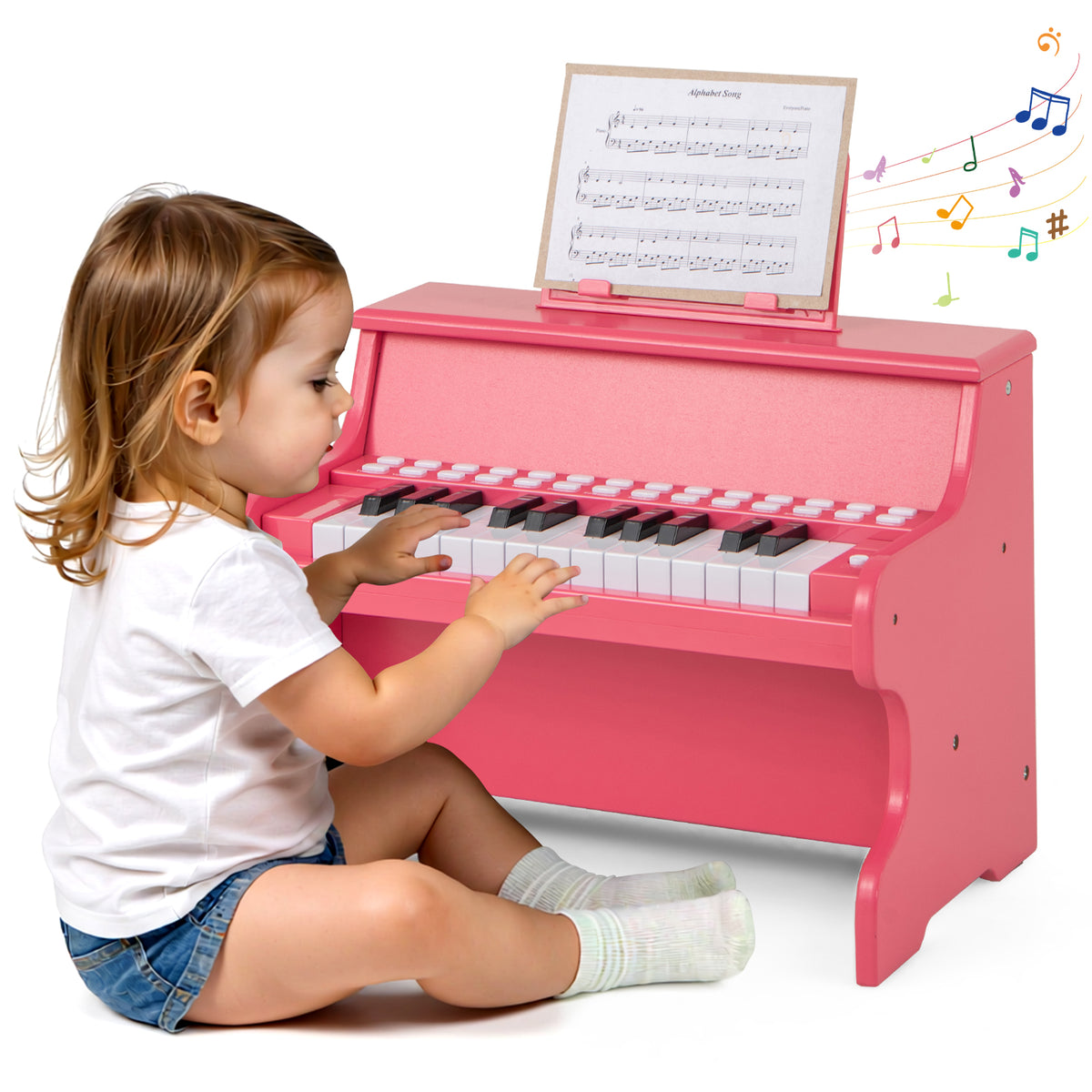 A toddler sits on the floor playing the pink wooden kids piano keyboard with sheet music