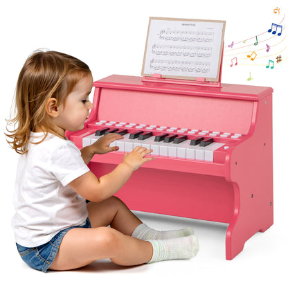 A toddler sits on the floor playing the pink wooden kids piano keyboard with sheet music