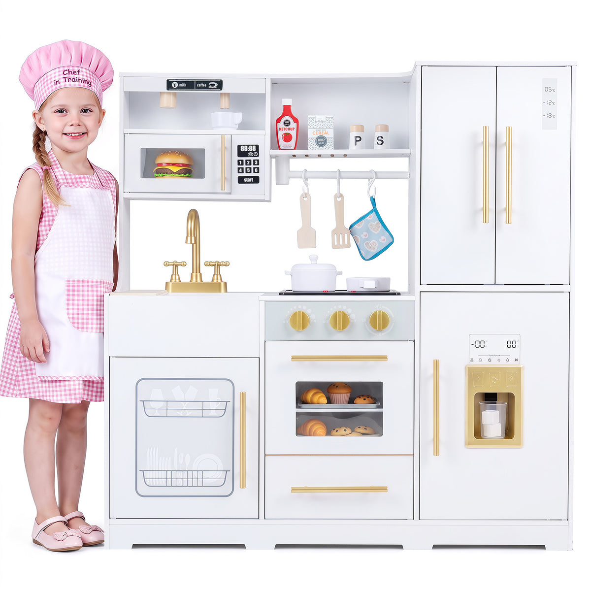 A young girl in a chef hat and apron stands by the white wooden play kitchen